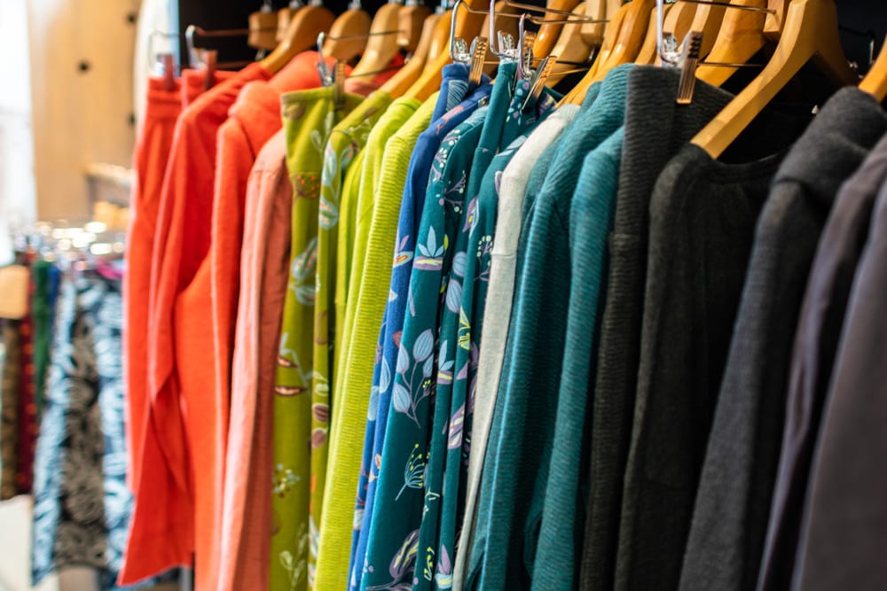 Das Angebot an Damenmode ist im Weltladen Klagenfurt nach Farben sortiert. Auf der Kleiderstange hängen Ober- und Unterteile von rot, orange, grün, blau, türkis bis schwarz. Die Kleiderbügel sind aus hellem Holz 