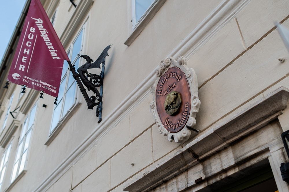 Die Außenansicht des Antiquariats in den Bärenlauben. Man sieht das alte Schild über der Tür mit Aufschrift auf Bronze, in der Mitte ist ein Relief von einem Baum in Gold, umrahmt ist das Schild mit einer Zierleiste in weiß. Links daneben hängt eine Fahne, gehalten von einem Drachen aus Eisen. Auf der roten Fahne steht "Antiquariat Bücher" sowie die E-Mail Adresse und Telefonnummer 