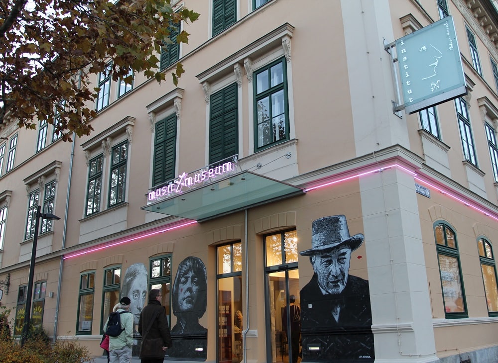 Das Musil-Haus von außen gegenüber vom Bahnhof. Im Erdgeschoss kommt man ins Musil-Museum, das steht in Leuchtschrift über den überdachten Eingang. Weiters zieren die Portraits von Robert Musil, Ingeborg Bachmann und Christine Lavant die Außenwand.