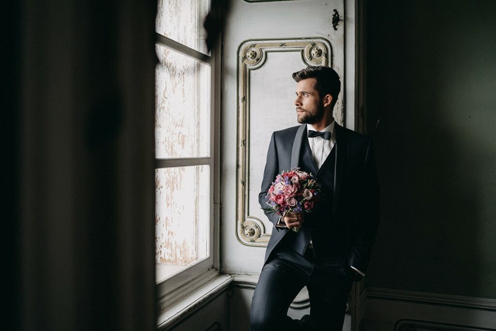 das perfekte Makeup für den Hochzeitstag auch für den Bräutigam bei Belinda Wrann art of beauty in Klagenfurt, das Bild zeigt einen Bräutigam im Hochzeitsanzug mit einem Brautstrauß, er steht an einem Fenster in einem Gang