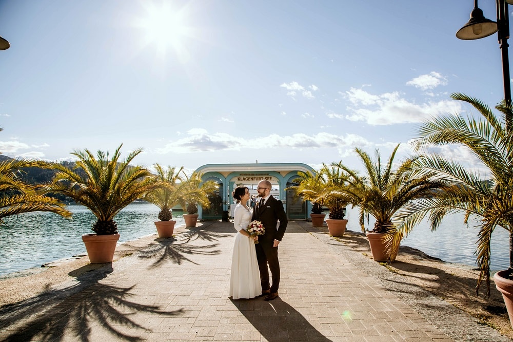 Ein Brautpaar an der Schiffanlegestelle in Klagenfurt am Wörthersee, sie trägt ein bodenlanges Brautkleid, er einen dunklen Anzug. Die Anlegestelle ist mit Palmen dekoriert, der Himmel ist strahlend Blau und die Sonne scheint, ein perfekter Hochzeitstag