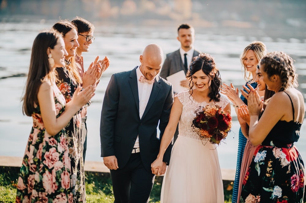 ein frisch verheiratetes Brautpaar schreitet gemeinsam durch das Spalier ihrer Freunde, die Hochzeit ist open Air, im Hintergrund der Wörthersee