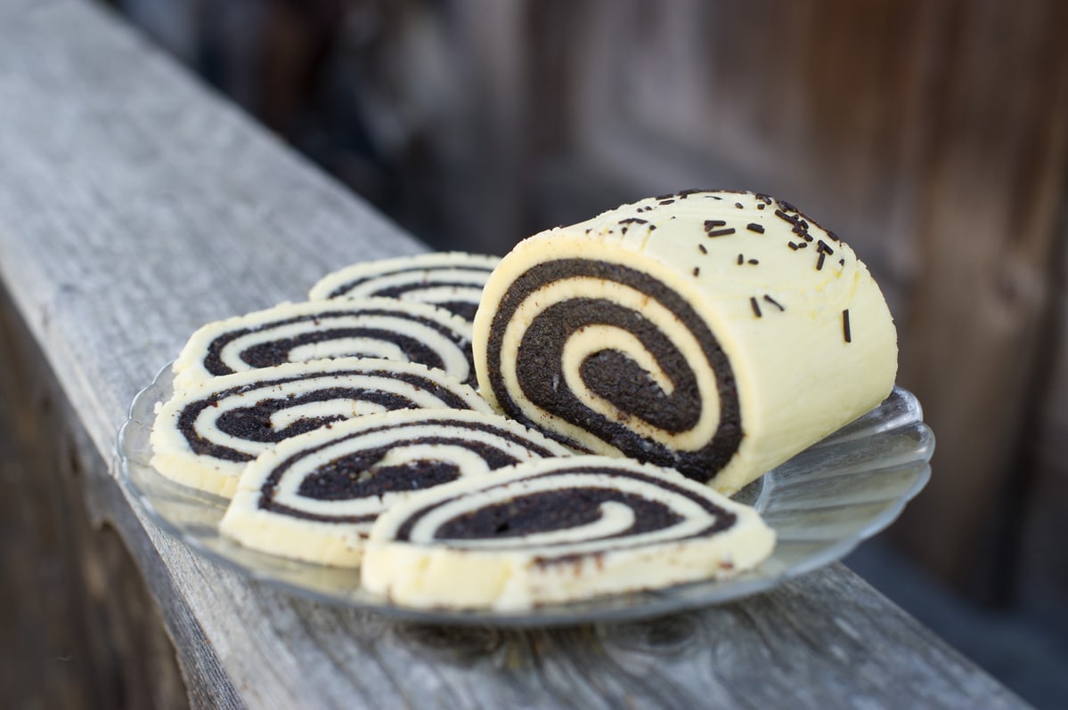 Mohnbutter in Schneckenform aufgerollt wurde in Scheiben geschnitten. Die Scheiben liegen auf einem durchsichtigen Teller auf einem Holzbalken. Das Restliche Stück liegt neben den Scheiben, man sieht den Anschnitt.