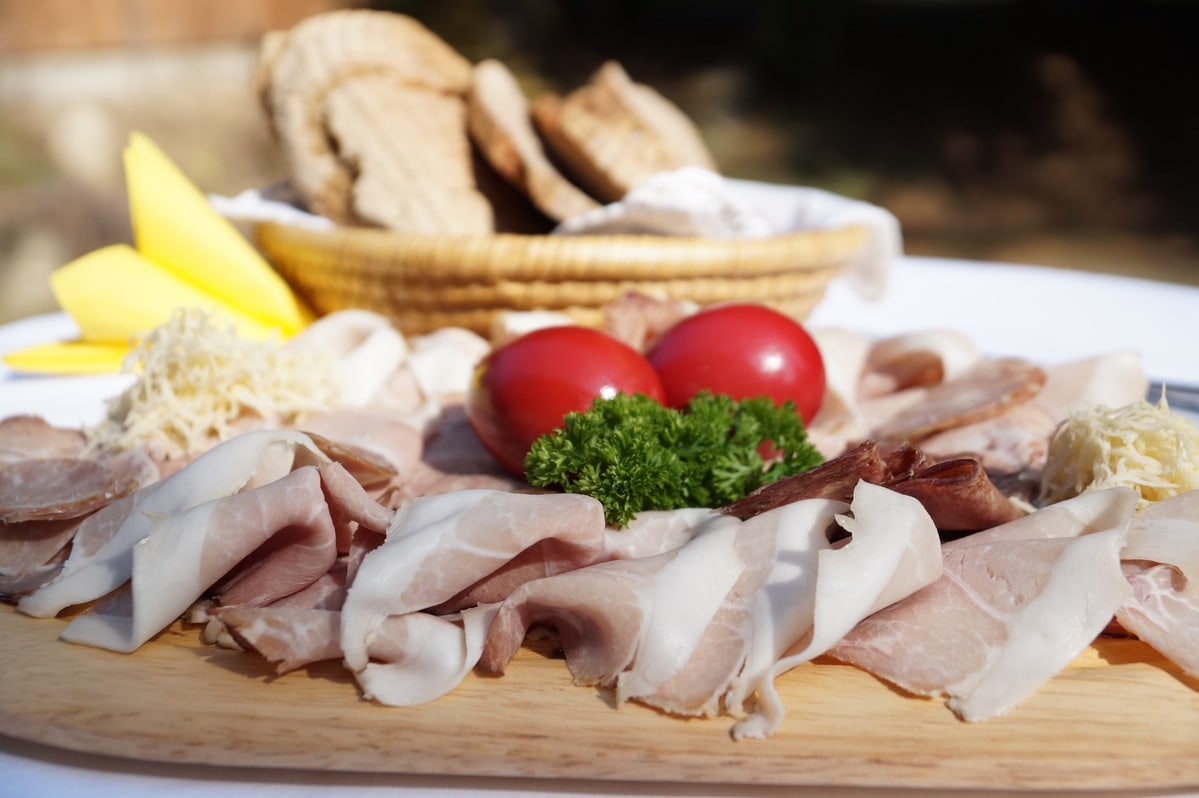 Auf einem Holzbrett ist aufgeschnitterner Schinken und andere Wurstwaren. Als Dekoration ist mittig Petersielie und zwei rote Ostereier platziert, rechts daneben ist geriebener Kren. Im Hintergrund ist ein Körbchen mit Brot. Alles steht auf einem Tisch mit weißer Tischdecke