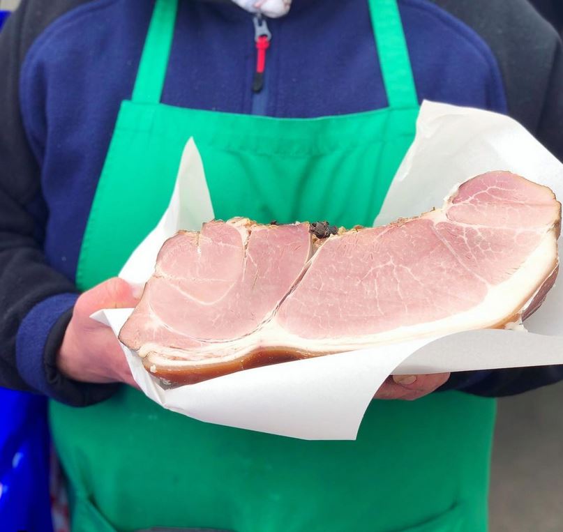 Am Bild sieht man den Oberkörper einer Person mit blauem Pullover und grüner Schürze. Mit beiden Händen hält die Person ein großes Stück Schinken, von dem man den Anschnitt sieht. Er liegt auf einem weißem Papier