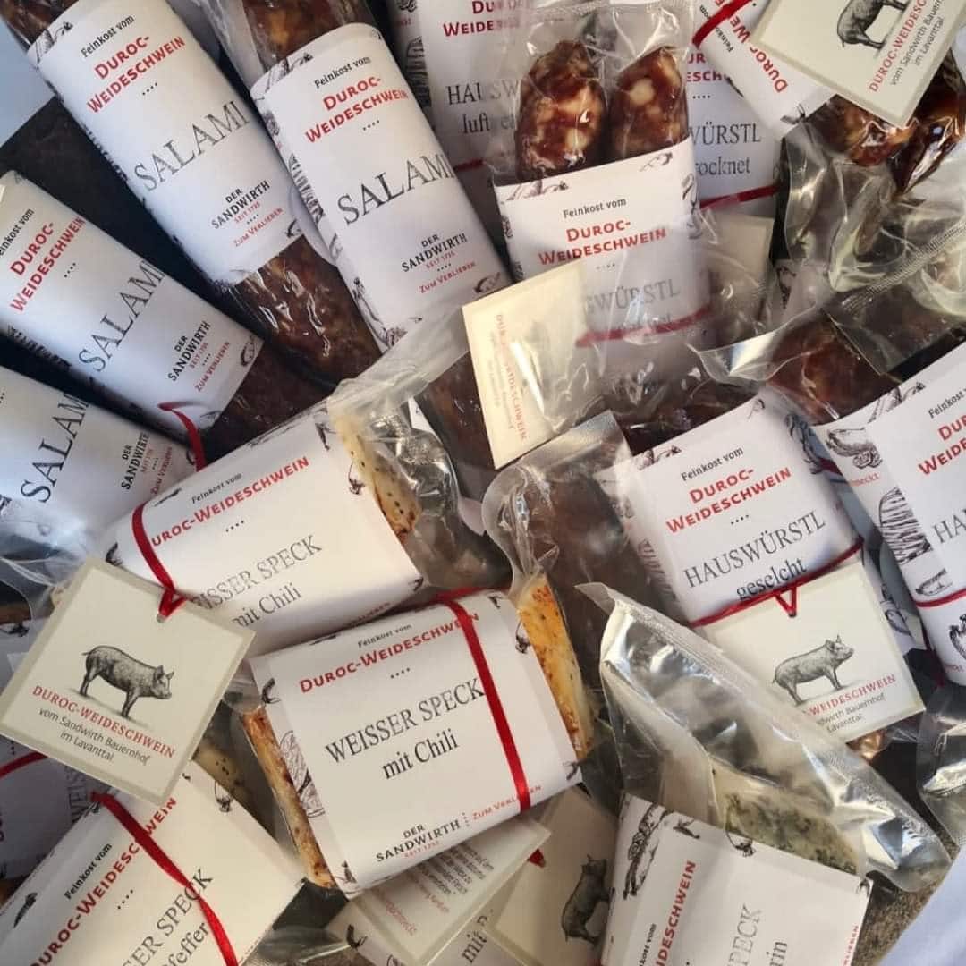 Von oben fotografiert sieht man viele in durchsichter Folie verpackte Fleischprodukte vom Sandwirth. Auf einem weißen Etikett auf jedem Produkt wird der Inhalt beschrieben. Z.B.: Hauswürstl, Speck, Salami. Mit einem roten Band ist das Schild mit dem illustrierten Schwein angebunden. Das Fleisch stammt vom Duroc-Weideschwein vom Sandwirth Bauernhof im Lavanttal.