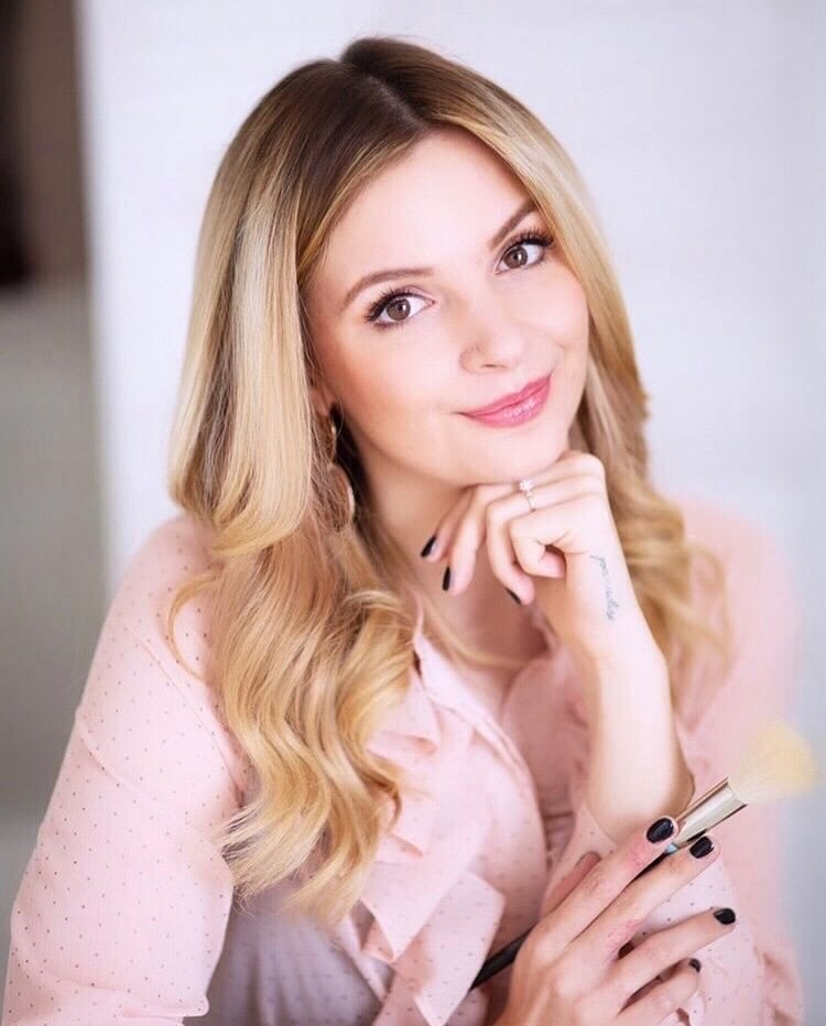 Blonde Frau mit langen Haare und rosa Bluse blickt in die Kamera, in der rechten Hand hlät sie einen Pinsel, der auf ihren Beruf als Kosmetikerin und Make Up Artisthinweist