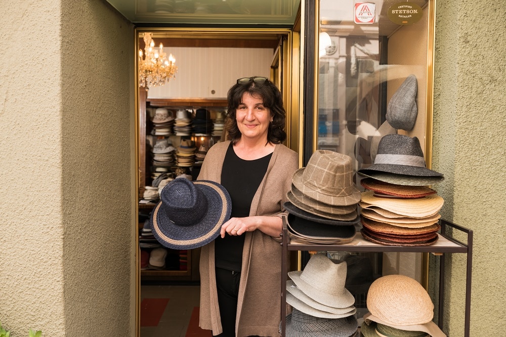 die Inhaberin des Geschäftes Modellhüte Luise lehnt am Türranhmen ihres Geschäfts in der Hand einen schwarzen Strohhut mit breiter Krempe und einem beigen Streifen, neben ihr ein Regal mit vier Stapeln mit Hüten aus unterschiedlichem Material