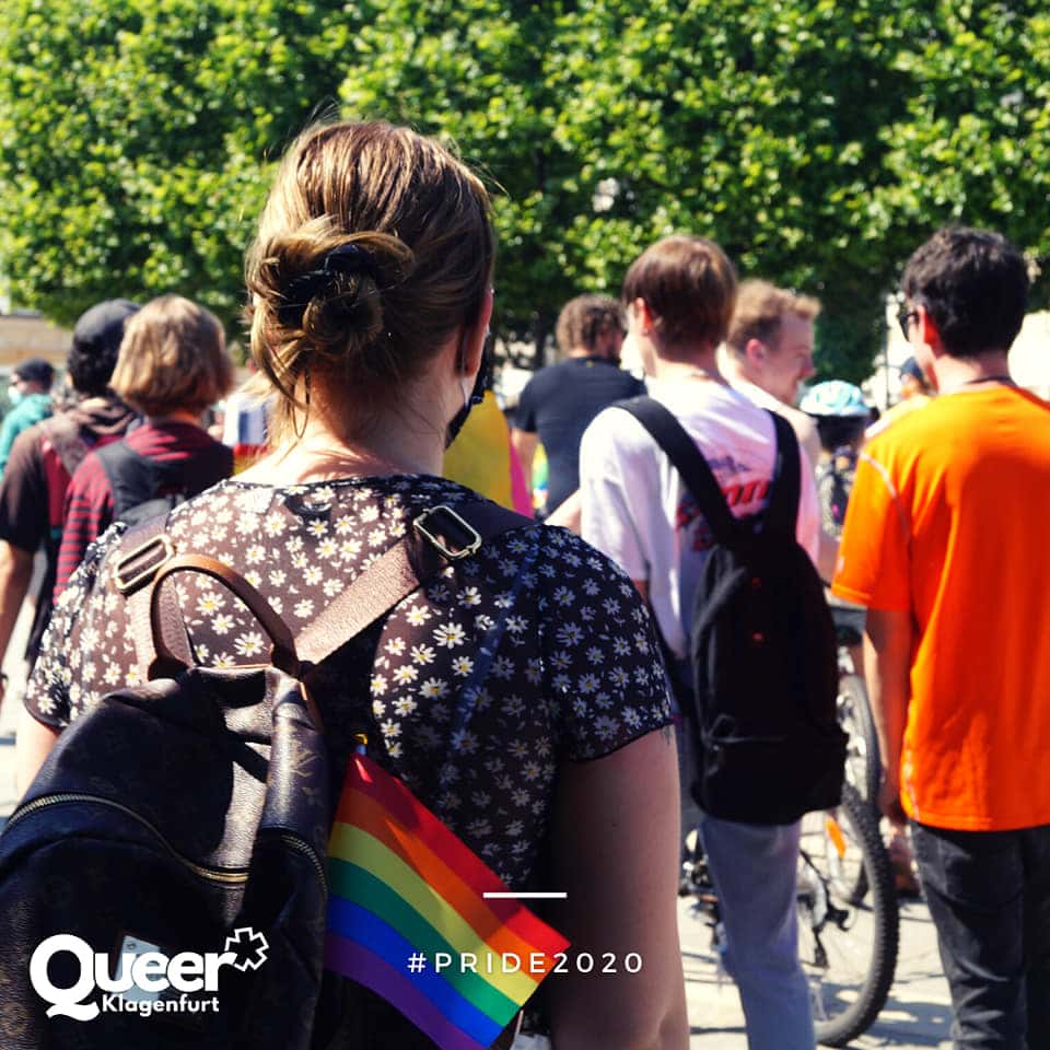 Rückansicht einer Frau mit Rucksack in dem eine Regenbogenfahne steckt, davor einige andre Menschen