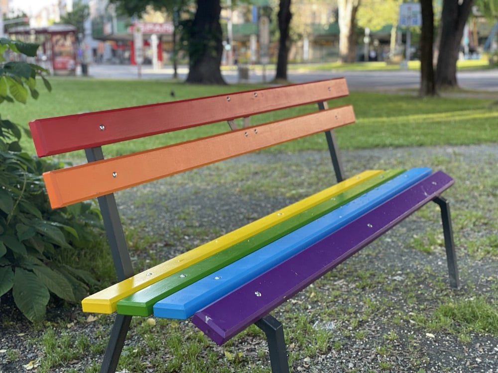 Parkbank in Regenbogenfarben vor der Kelag Klagenfurt, im Hintergrund Bäume und Sträucher