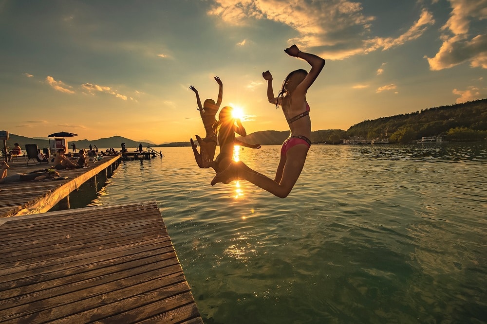 Abendstimmung im Strandbad Klagenfurt, die Sonne steht tief, man sieht einen Abschnitt der Brücke, auf dem Menschen liegen, im Vordergrund springen drei Mädchen mit erhobenen Händen ins Wasser