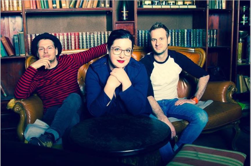 Die Band Acoustica bestehend aus Frontfrau Angelika und den Herren Roman und Benjamin sitzen auf einer Couch vor einer Bücherwand, Angelika trägt roten Lippenstift und eine blaue Bluse, Roman sitzt links im Bild in einer lässigen Haltung mit der Hand am Kinn und überschlagenen Beinen, er trägt einen Hut, Benjamin sitzt rechts im Bild mit in Jeans und einem Baseballshirt in schwarz-weiß