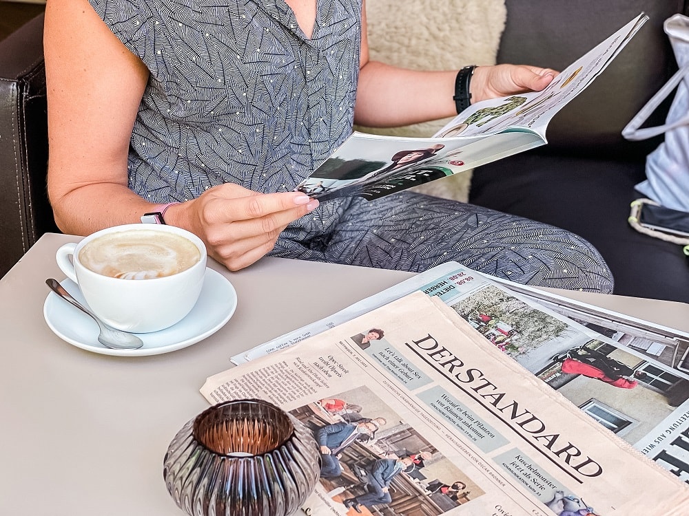 Frauenhände halten ein Magazin, auf dem Tisch liegen Tageszeitungen, daneben steht eine große Schale mit Kaffee und Milchschaum