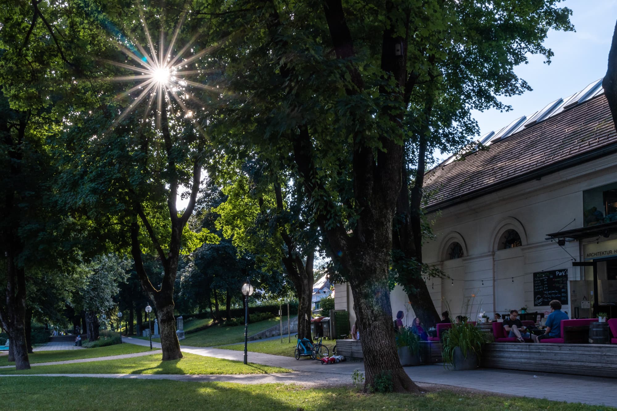 Sommerabend im Parkhaus Göthepark, die Sonne blinzelt durch die grünen Baumkronen, links genießen noch einige Gäste den lauen Sommerabend im Parkhaus Klagenfurt