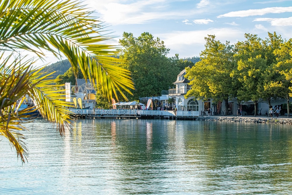 Blick auf die Villa Lido, im Vordergrund eine Palme und er blaue Wörthersee, ein Schiff legt an