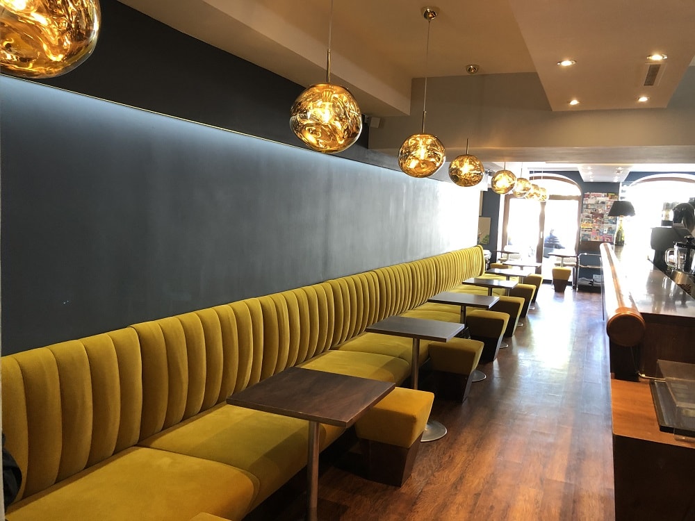 Blick ins Café am Platz mit einer durchgehenden, an der Wand verbauten Couch aus gelbem Samt, dazu kleine, eckige Bistrotische und goldene, kugelförmige Lampen, am rechten Bildrand ist die Theke zu sehen
