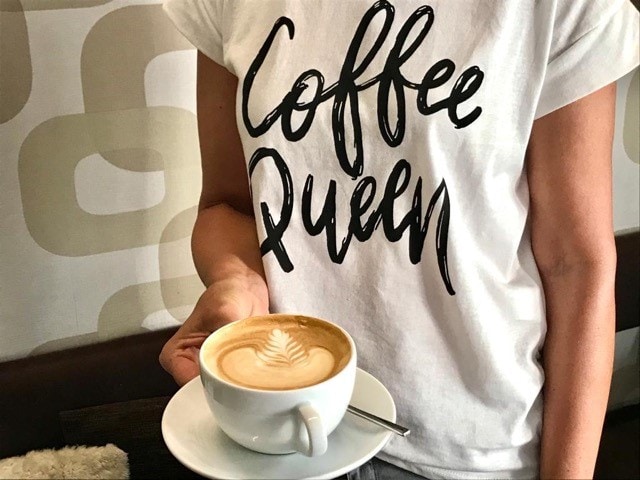 der Bildausschnitt zeigt die Arme und das T-Shirt einer Frau, die einen Cappuccino mit Milchschaum in der Hand hält, auf den ein Blatt gezeichnet ist, auf ihrem T-Shirt steht coffee queen