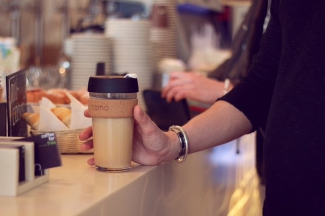 eine Hand mit Armreif stellt einen COMO-To-GO-Becher auf den Tresen