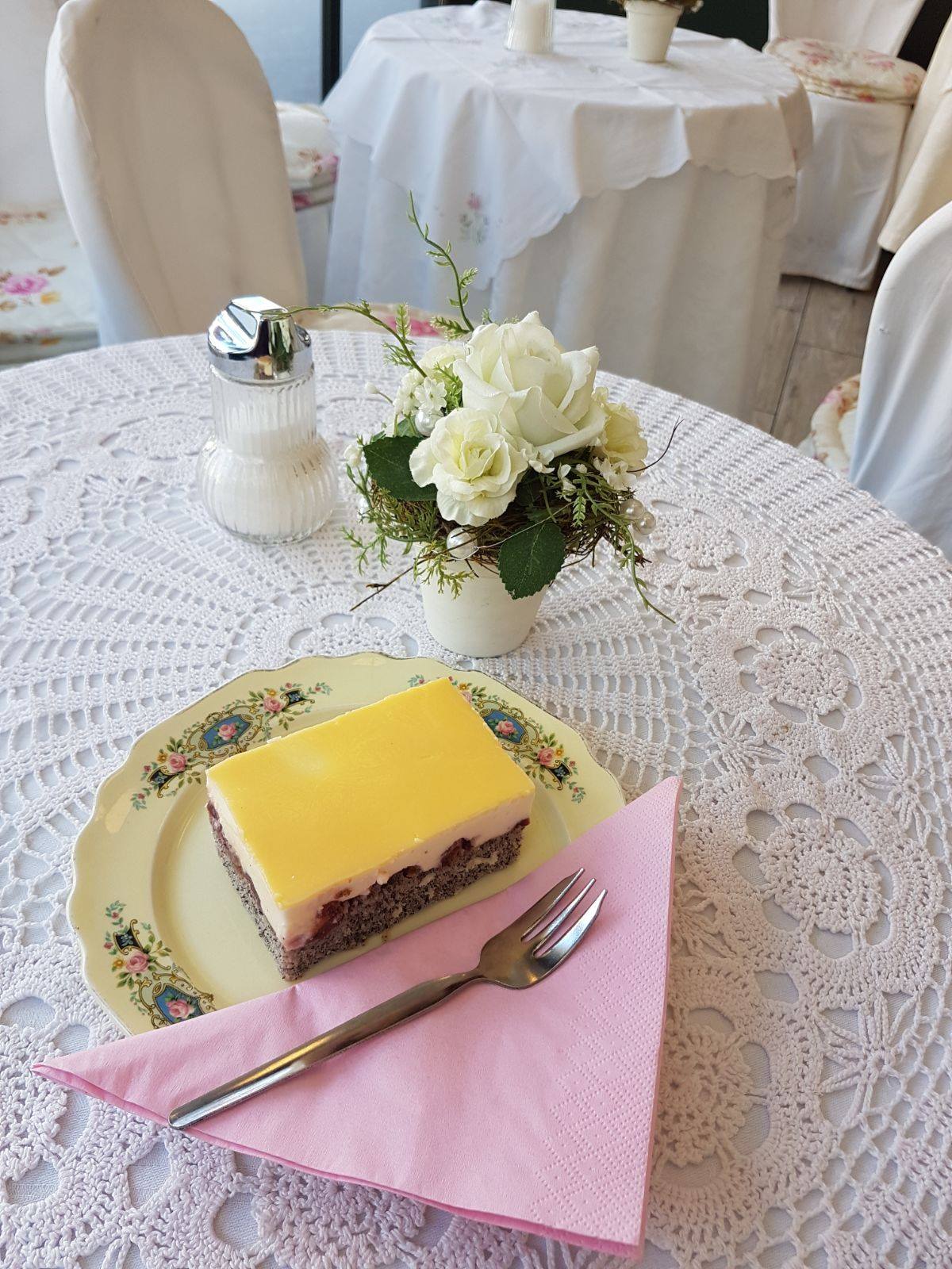 die beliebte und hausgemachte Eierlikörschnitte von Sylvies Kostbarkeiten am Benediktinermarkt in Klagenfurt. Liebevoll angerichtet auf einem verzierten Porzellanteller mit rosa Serviette. Der Tisch ist mit einem Häkeldeckchen und einen kleinen Topf mit einer weißen Rose geschmückt