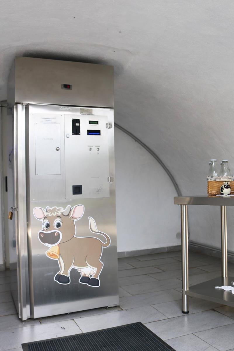 der 24 Stunden Frischmilchautomat am Hoislhof aus Edelstahl, daneben stehen Milchflaschen aus Glas, am Automaten klebt ein Aufkleber einer Comic-Kuhl mit goldener Glocke