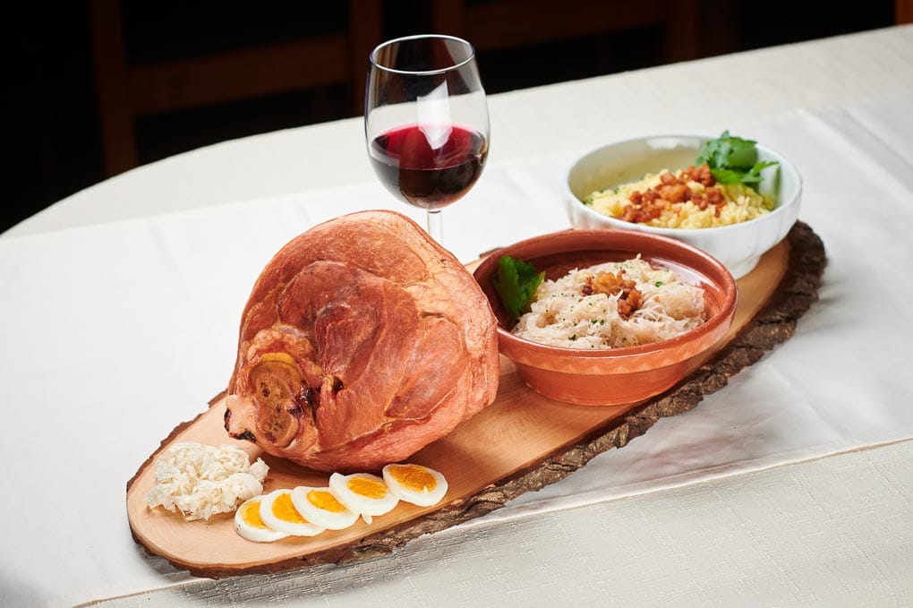 Stelze, Kren, Ei, Krautsalat und Rösti mit einem Glas Rotwein auf einer Holzplatte 
