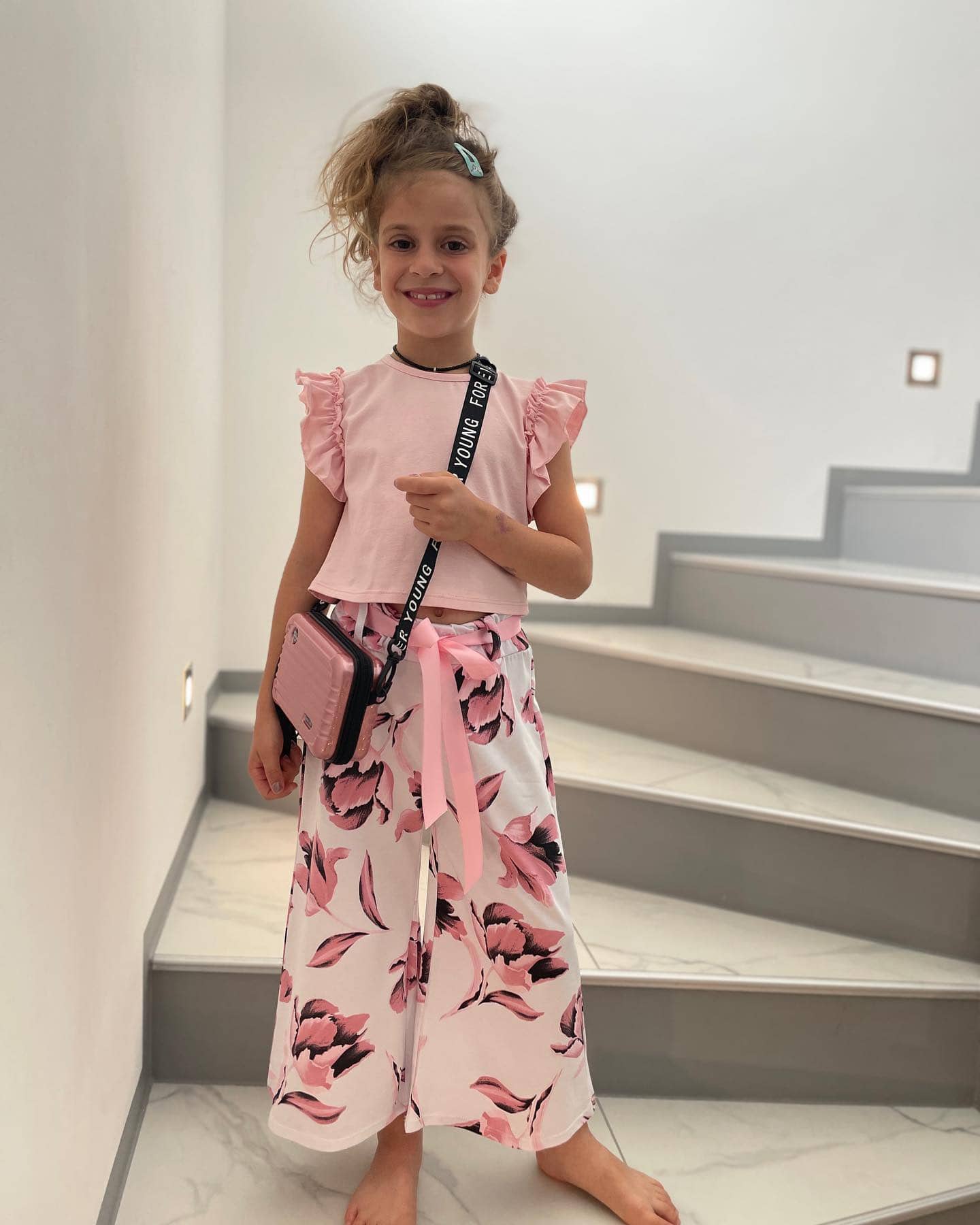 Mädchenoutfit zum Schulbeginn in Rosa mit einfarbigem Shirt mit Flügelärmeln und einer weit ausgestellten Hose mit Blüten, dazu eine rosa Umhängetasche