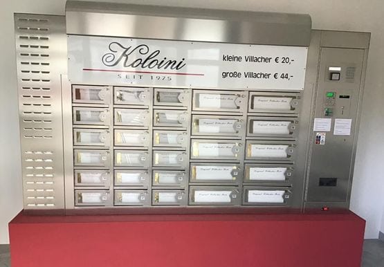 Tortenautomat mit verschiedenen Fächern aus Metall mit Beschriftung, welche Torte sich darin befindet, die Aufschrift verrät, dass die kleine Villachertorte 22€ und die große 44€ kostet, auf der rechten Seite der Box ist ein Münzeinwurf und ein Kartenterminal zu sehen