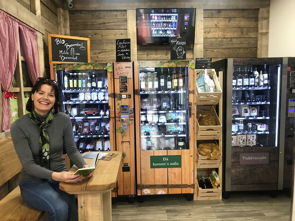 24h Automatenhäuschen mit drei Vitrinen, gefüllt mit regionalen Produkten aus Kärnten wie Milch, Öl, Kekse, Speck, Schinken, viele Bioprodukte, eine freundlich lächelnde Frau, die auf einer kleinen, hölzernen Eckbank sitzt und in einerm Buch blättert
