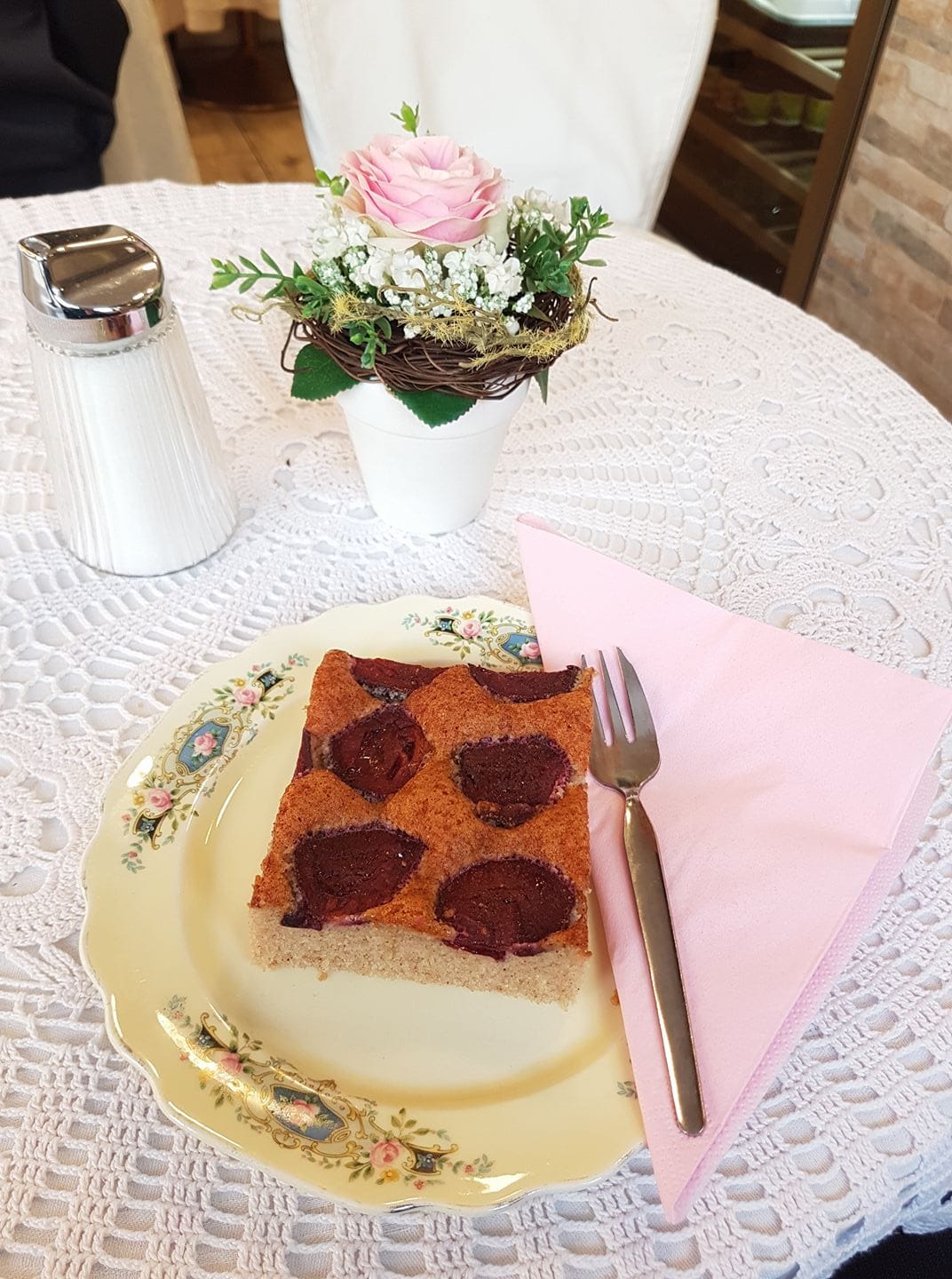 der beliebte und hausgemachte Zwetschkenkuchen von Sylvies Kostbarkeiten am Benediktinermarkt in Klagenfurt. Liebevoll angerichtet auf einem verzierten Porzellanteller mit rosa Serviette. Der Tisch ist mit einem Häkeldeckchen und einen kleinen Topf mit einer rosa Rose geschmückt