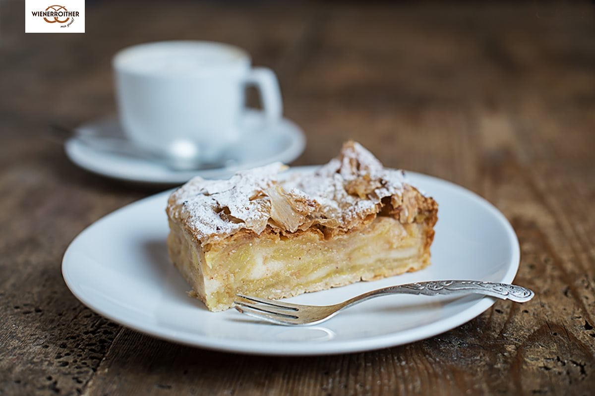 ein hausgemachter Apfelstrudel von der Bäckerei Wienerroither, serviert auf einem weißen Teller mit einer silbernen Kuchengabel. im Hintergrund eine Kaffeetasse