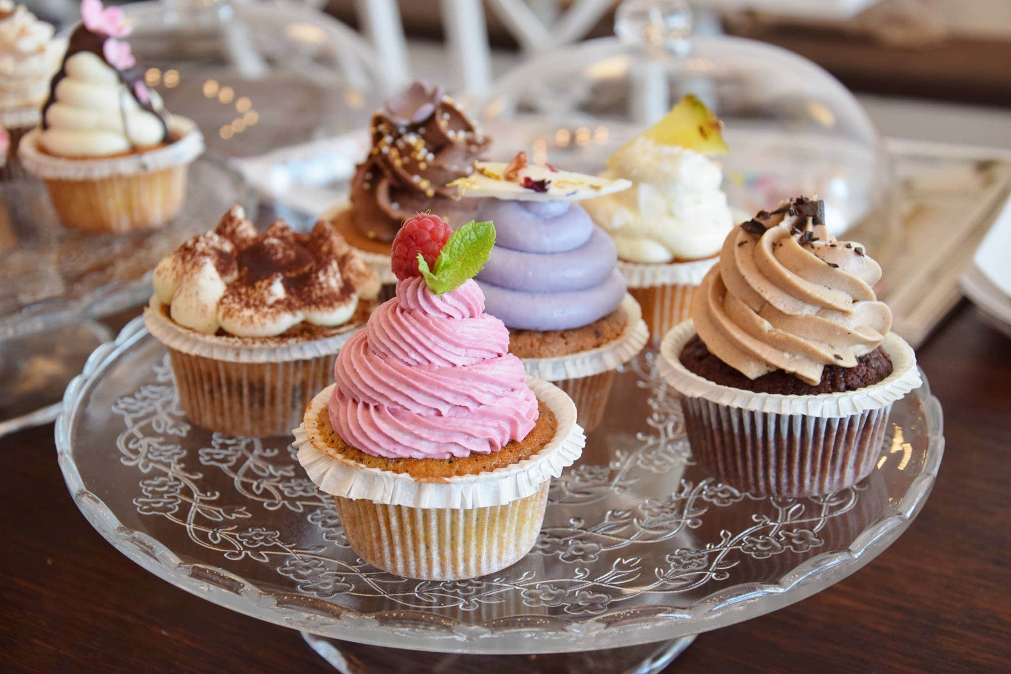 ein Glasteller mit verschiedenen Cupcakes von Rose Cupcake Shop & Cafe in Klagenfurt. Sie haben unterschiedlichen Kuchenteig und verschiedenfarbige Toppings verziert mit Beeren, Schokolade, Streusel und Kakao