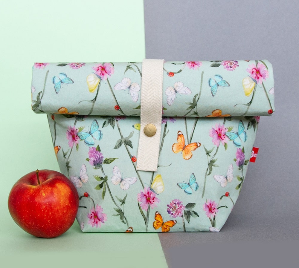 ein mintfarbener Lunchbag mit Blumen und Schmetterlingsaufdruck und einem Verschlusss mit Druckknopf, Design aus Klagenfurt von Veinzelstück, daneben ein Apfel