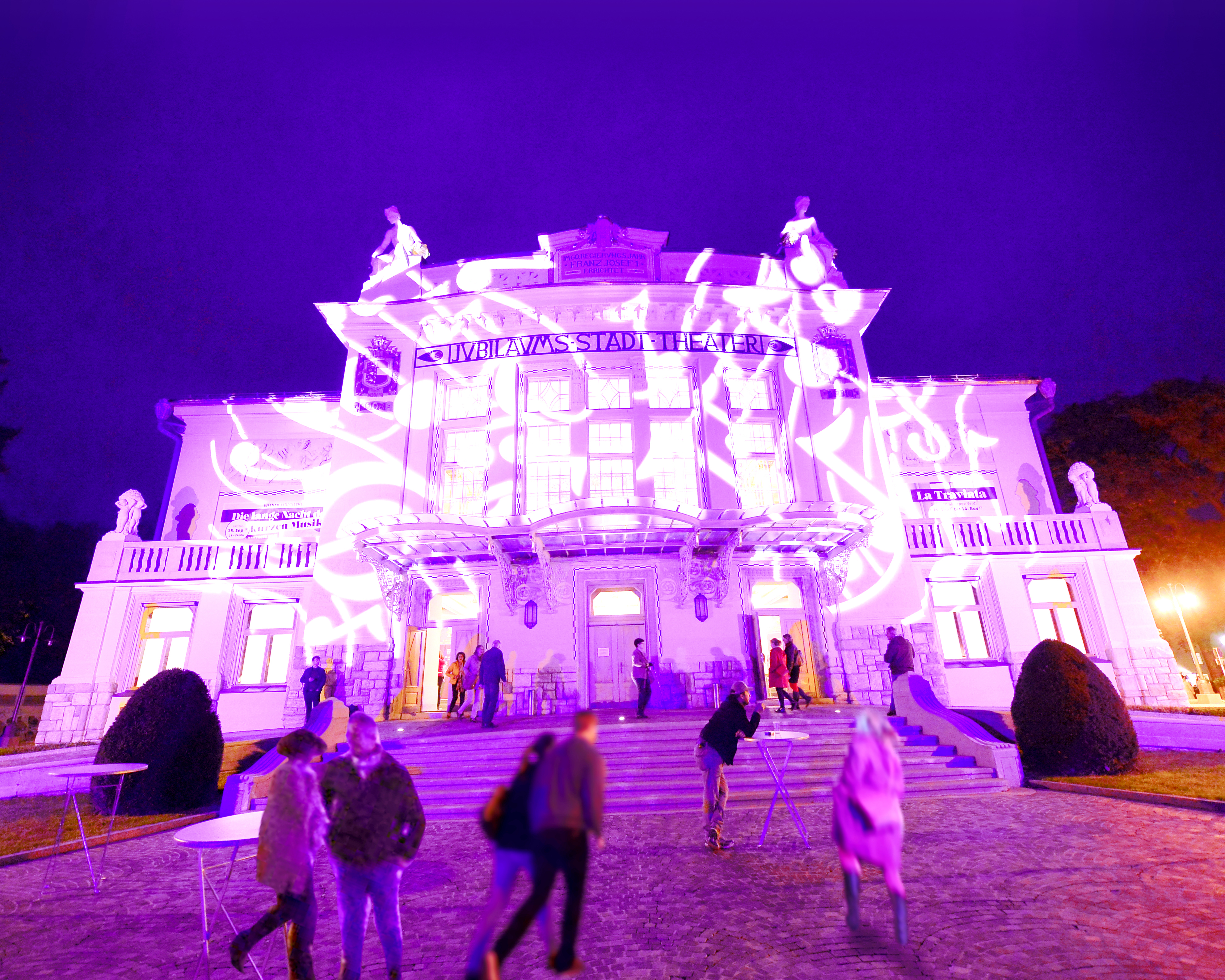 Stadttheater Klagenfurt in Lila, bei Dunkelheit, Personen am Theaterplatz