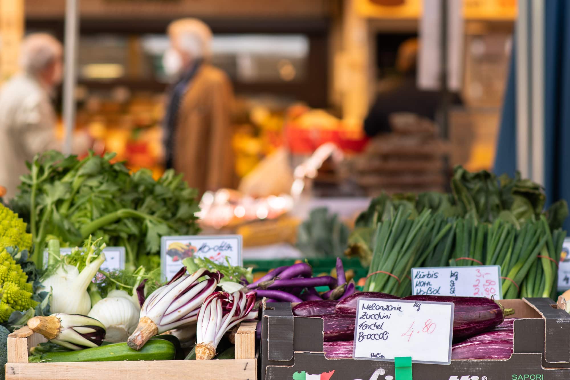 Gemüsestand am Benediktinermarkt - im Vordergrund erkennt man verschiedenes Gemüse wie Salate und Jungzwiebeln in Kisten und den Preisschildern. Im Hintergrund ist unscharf ein weiterer Marktstand zu erkennen, auf dem vor allem gelbes und oranges Obst und Gemüse präsentiert wird. Davor ist ein Mann mit hellbrauner Jacke, grauem Schal, weißer FFB2-Maske und weißen kurzen Haaren zu sehen, welcher sich, relativ mittig im Bild, nach links gedreht vorbeigeht. Ebenso ist am rechten Bildrand eine weitere Person mit braunen Haaren und schwarzer Jacke von hinten zu sehen. Von links kommt eine Dame mit weißer Jacke, weißen kurzen Haaren und FFB2-Maske ins Bild hinein.