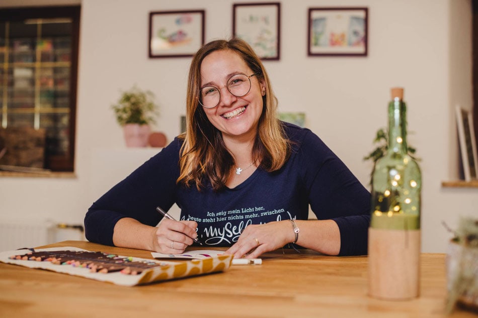 die Glückskleckserin Melanie Riemer sitzt an ihrem Tisch mit einem Stifteset und schreibt, dabei lächelt sie in die Kamera