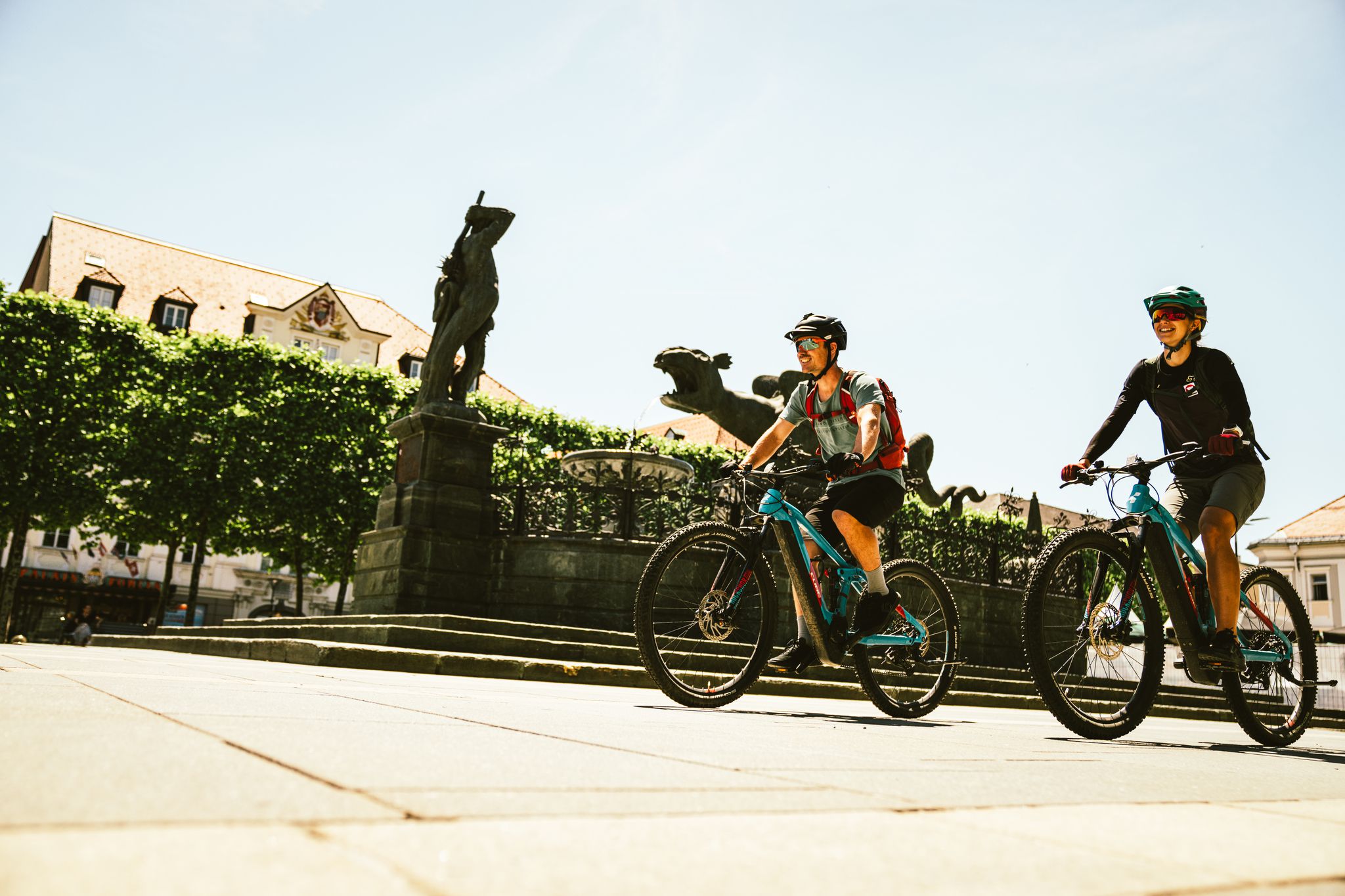 2 Radfahrer fahren mit ihren Rädern am Lindwurm in Klagenfurt vorbei