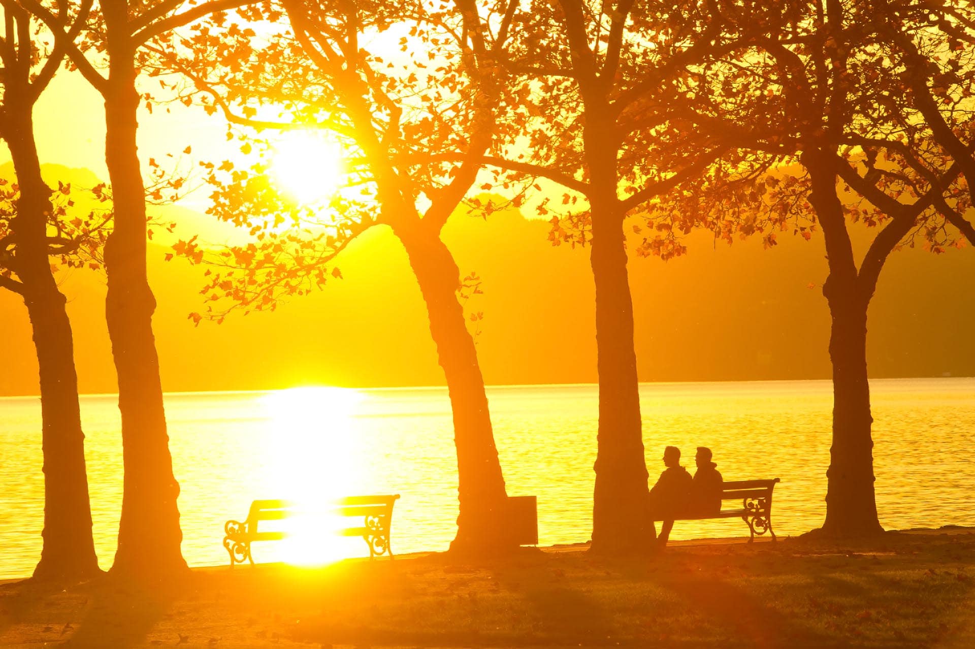 2 Personen sitzen auf Parkbank und genießen den Blick auf den Wörthersee bei Sonnenuntergang 
