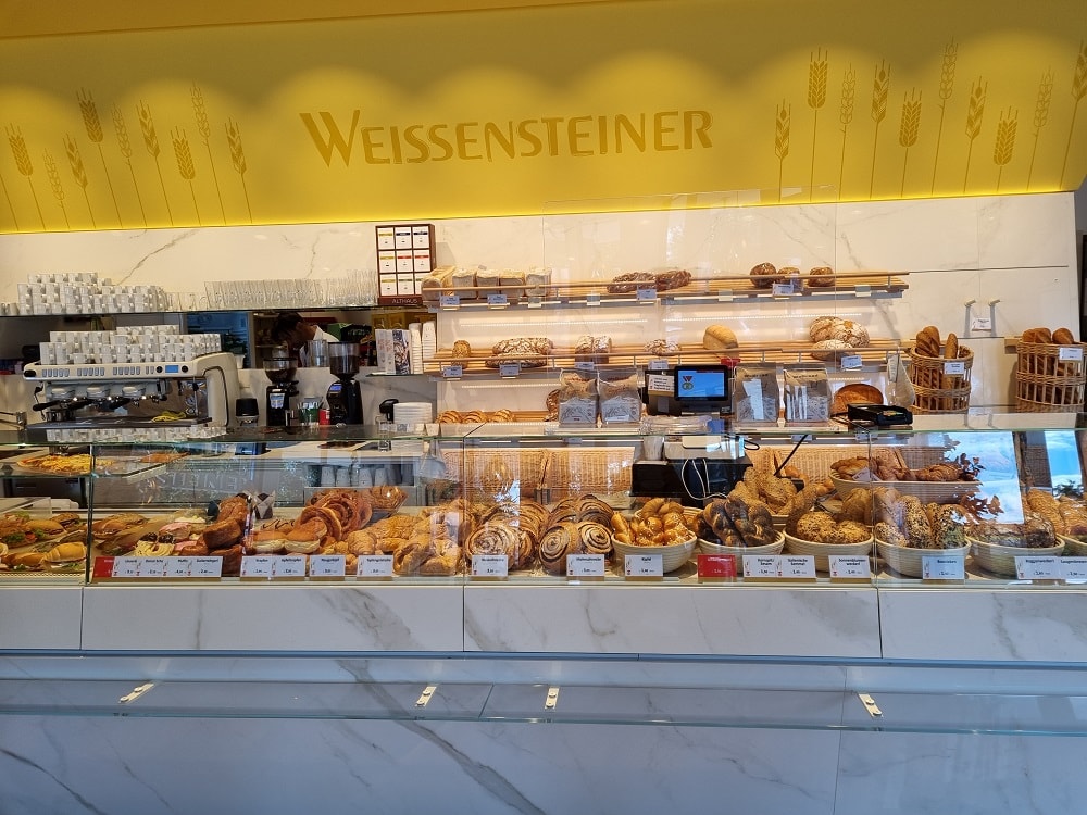 Innenansicht der Bäckerei Weissensteiner mit einer Glasfront hinter der viele verschiedene Backwaren zu sehen sind: Nusschnecken, Mohnzöpfe, Kornspitz, Brot, Semmeln, Krapfen, Kornsemmeln und eine Kaffeemaschine