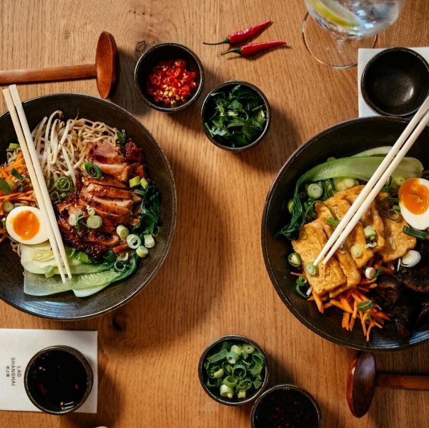 Ein Menü mit zwei Tellern von Lao Shanghai, links gebratene Ente mit Nudeln, Gemüse und Ei, rechts gebratener Tofu mit Gemüse und Ei, dazu ein paar Gewürze und Stäbchen