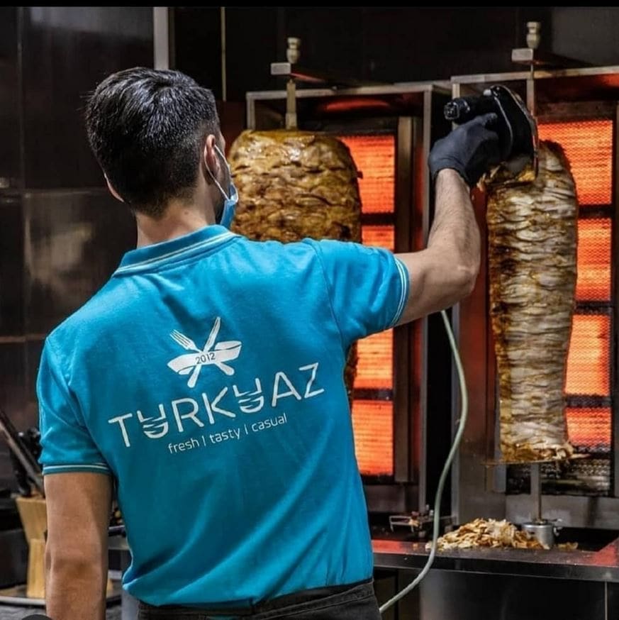 der Mitarbeiter bei turkuaz in den City Arkaden steht vor einem Dönerspieß und schneidet Fleisch, er trägt ein T-Shirt mit dem Markenlogo