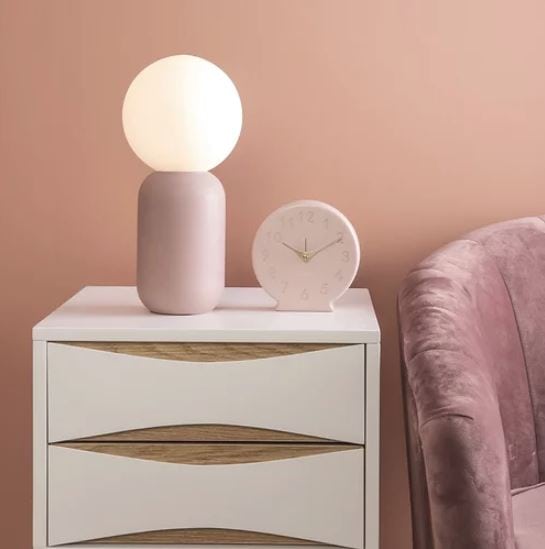 Table Lamp Gala ist eine Tischlampe in Form eines Zylinders mit einer leuchtenden Kugel darauf. Am Bild steht die Lampe auf einem weißen Kasterl mit Laden neben einer rosafarbenen Couch. Neben der Lampe steht ein weißer Wecker. 