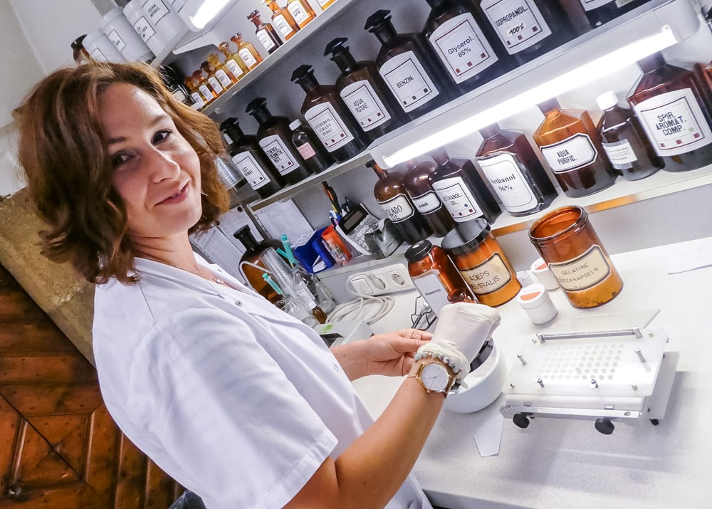 In einem hinterem Raum in der Engel Apotheke werden die Eigenprodukte hergestellt. Am Bild ist eine junge Dame, die eine Salbe anmischt. Sie hat dunkle, schulterlange Haare, einen weißen Kittel und trägt an der rechten Hand einen Einweghandschuh. Auf den Regalen rechts über den Tisch sind die verschiedenen Zutaten in unterschiedlich großen Glasbehältern mit weißen Etiketten. Am Tisch steht eine Waage und offene Gefäße.