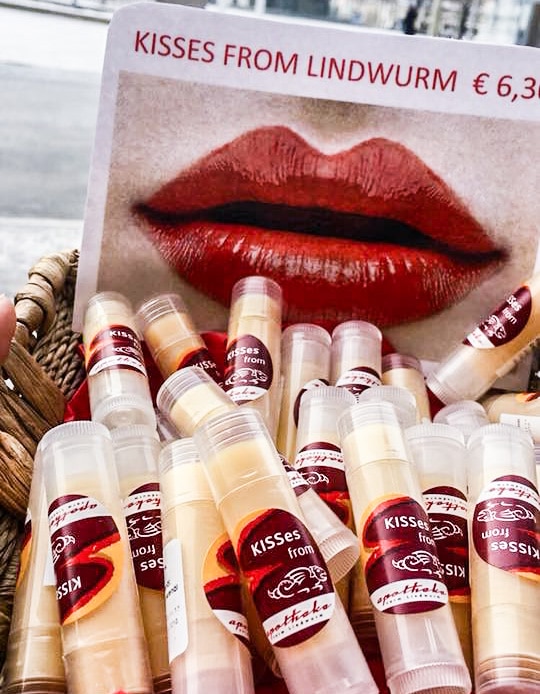 Als Lippenpflege bekommt man in der Apotheke vorm Lindwurm ein Eigenprodukt namens " KISSes from" mit Lindwurm darunter. In einem Korb sind mehrere Sticks aus durchsichtiger Plastikverpackung zu sehen. Das Etikett ist rot. Der Inhalt hellgelb. Im Korb ist ein Schild gesteckt, dass den Markennamen, den Preis und darunter groß eine rot geschminkte Lippe zeigt. Im Hintergrund ist aus dem Fenster hinaus leicht die Straße zu erkennen.