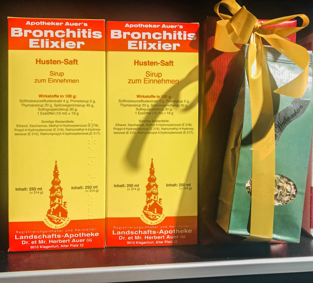 Die Landschaftsapotheke bietet einen Hustensaft aus Eigenproduktion an. Auf dem Bild sind zwei Packungen des "Bronchitis Elixiers" zu sehen. Die hochformatige Packung ist am oberen und unteren Ende rot mit weißer Schrift, der breitere mittlere Teil ist gelb mit roter und schwarzer Schrift. Das Logo unten stellt den Stadtpfarrturm dar. Er ist ebenfalls rot. Rechts neben den beiden Verpackungen des Hustensaftes steht eine Teemischung der Apotheke in grüner Tüte mit Sichtfenster und gelber Schleife.