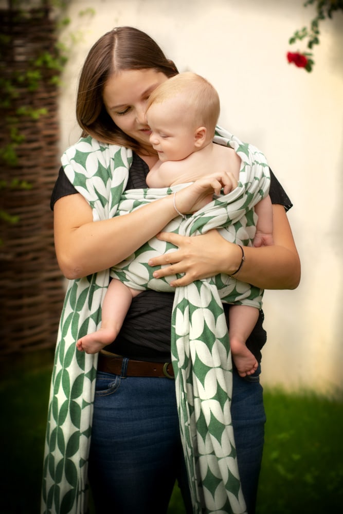 Am Bild ist eine Frau mit Baby im Arm zu sehen. Sie ist von vorne zu sehen und blickt auf das Baby, das sie in ihr weiß-grün gemustertes Tragetuch wickelt. Das Baby lächelt. Im Hintergrund ist unscharf eine grüne Wiese und eine weiße Wand zu erkennen. Von rechts oben hängt eine rote Rose ins Bild.