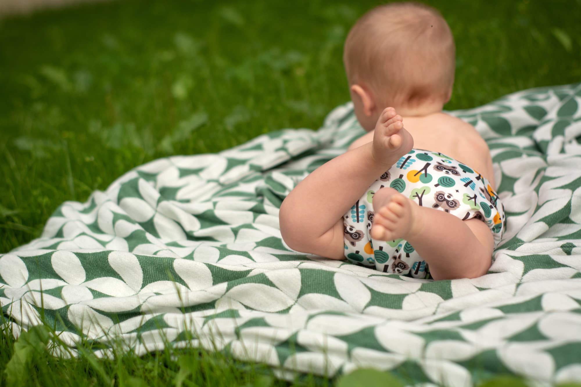 Ein Baby mit Stoffwindel liegt auf dem Bauch, den Kopf von der Kamera weggedreht, die Beine angewinkelt in der Luft. Auf der Stoffwindel sind auf weißem Hintergrund Illustrationen von Bäumen und Waschmären. Das Baby liegt auf einer Baumwolldecke mit weiß-grünem Muster, die auf einer grünen Wiese liegt.