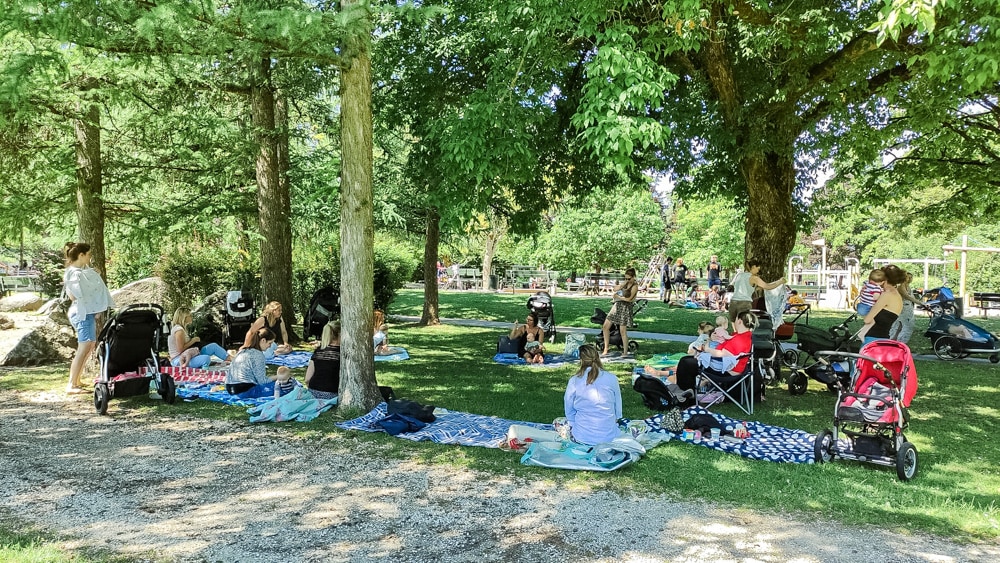 In einem Park an einem sonnigen Sommertag, im Hintergrund ist ein Spielplatz zu sehen, haben sich mehrere Mütter mit ihren Babys zur Stillgruppe getroffen. Es sind einige Decken am Boden im Schatten der großen Bäume ausgebreitet und es sind auch viele Kinderwägen zu sehen