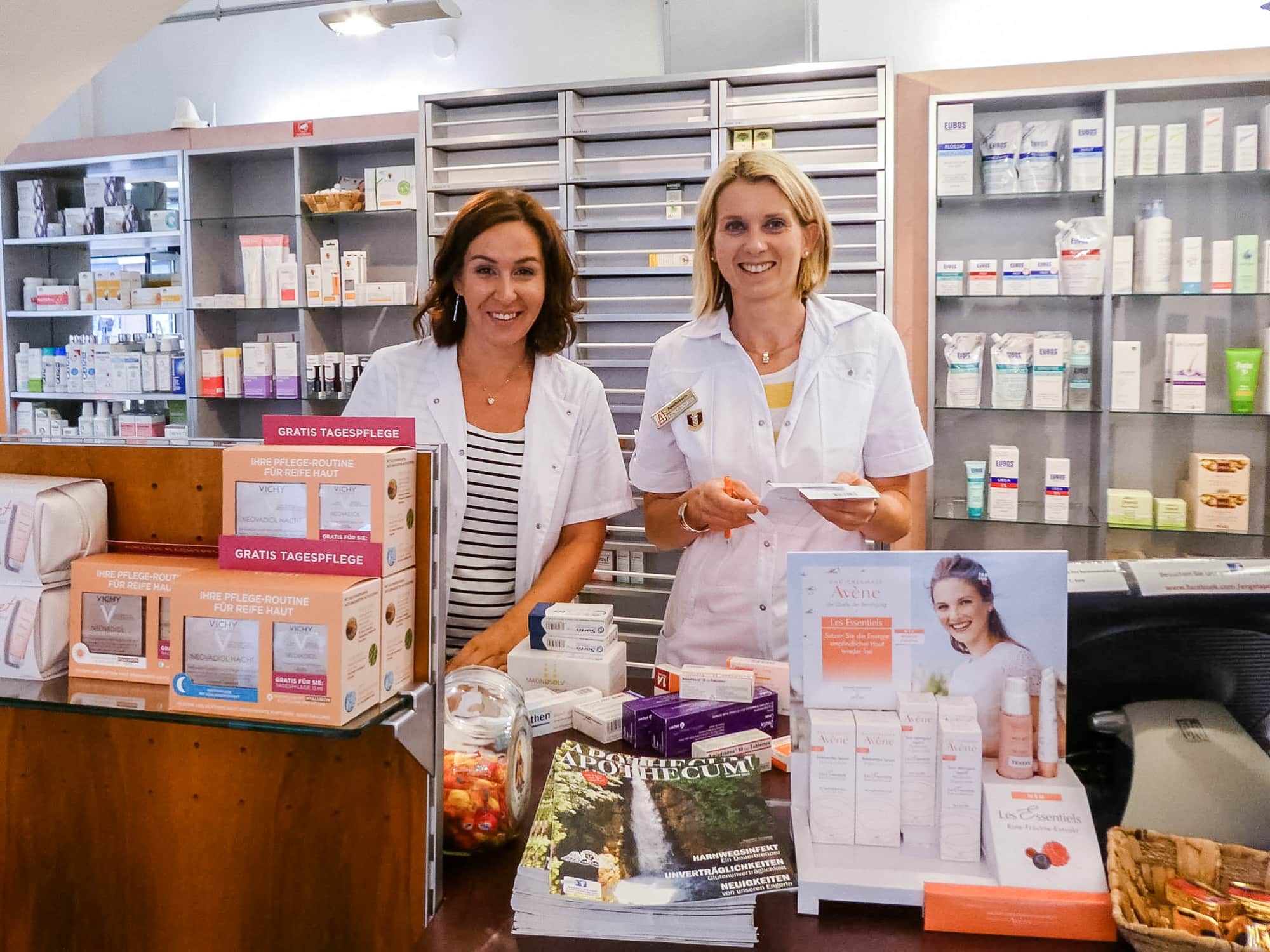 Zwei Damen der Engel Apotheke stehen hinter dem Verkaufstresen und lächeln in die Kamera. Am Tresen aus dunklem Holz liegen mittig Zeitschriften, dahinter und rechts daneben sind Produkte, links ist auf der Erhöhung mit Glasablage auf der ebenfalls Produkte stehen. Im Hintergrund sind Laden und Regale im hellen Design mit Pflegeprodukten.