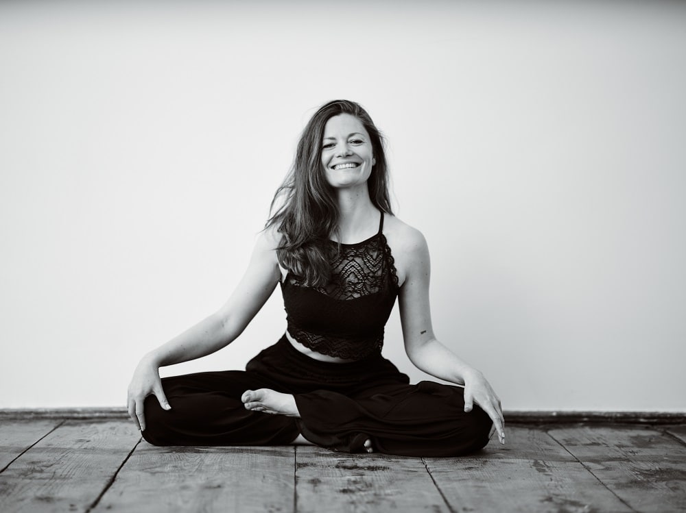 Yogalehrerin Patricia Leitgeb persönlich, sie sitzt im Yogasitz auf einem Holzboden an eine Wand gelehnt und trägt schwarze Yogakleidung, sie lächelt