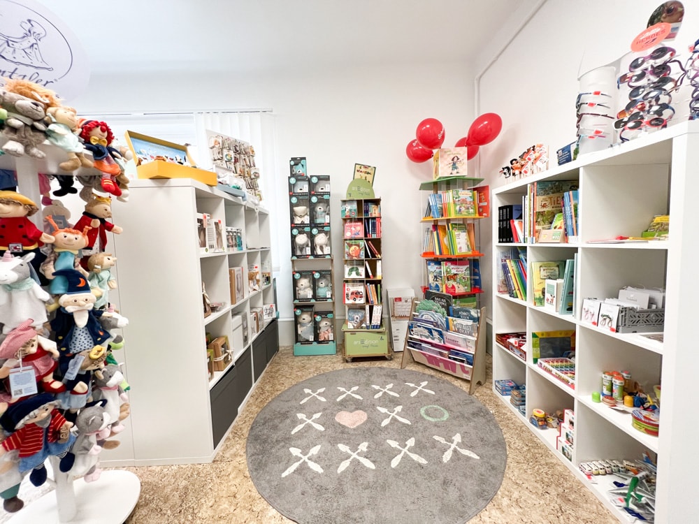 Am Bild ist ein Geschäft mit Kinder- und Baby-Spielzeugen zu sehen. Rechts an der Wand, in der Ecke und links am Bild in den Raum stehend sind weiße Regale und Ständer aufgestellt, die mit Büchern, Stofftieren und anderen Dingen befüllt sind. Zwischen den Regalen liegt ein runder grauer Teppich.